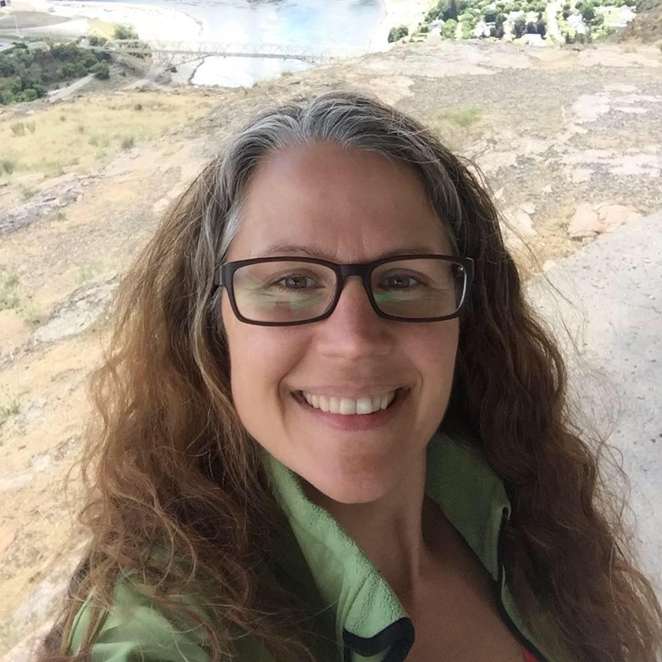 A woman with glasses is taking a selfie in front of a lake