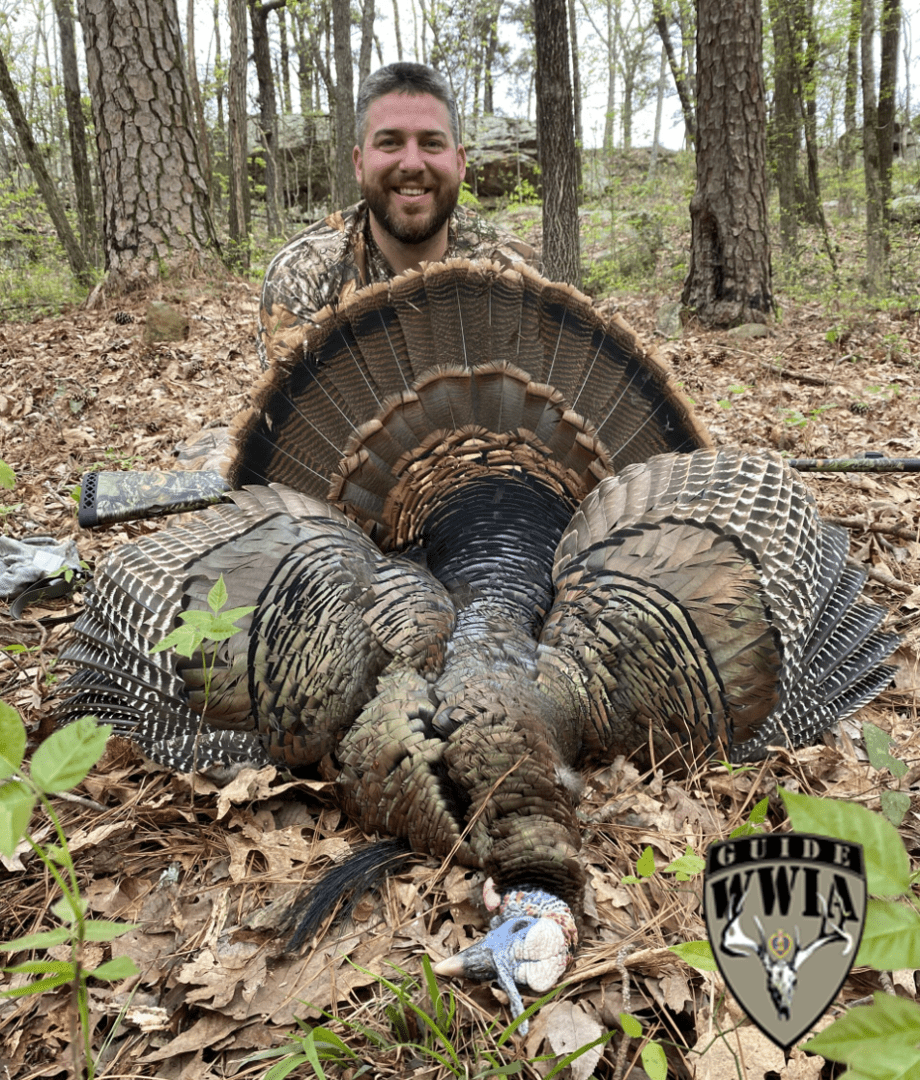 A man posing with a turkey in the woods.