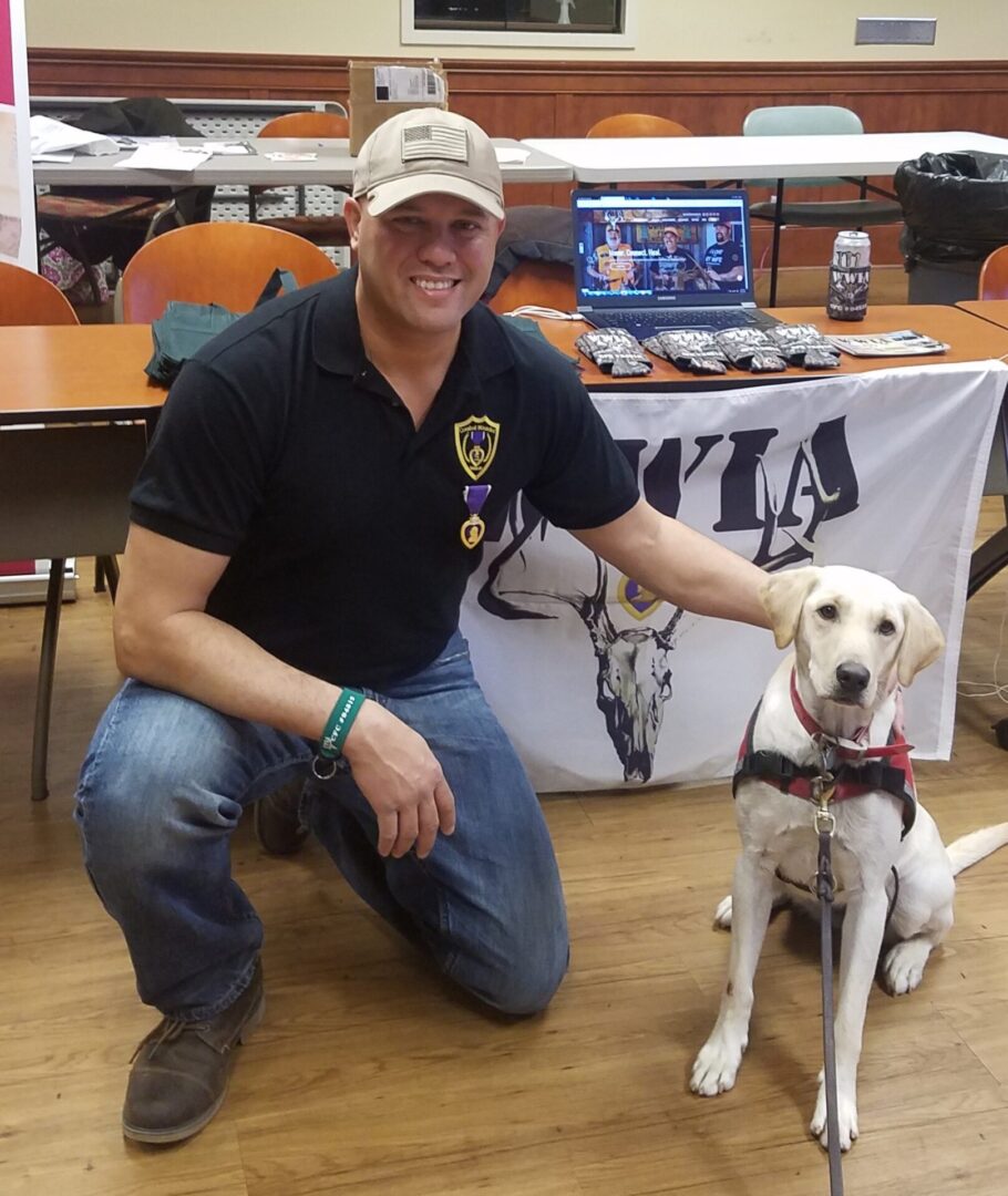 A man in a cap posing with a dog next to a table with military insignia and information displays at an indoor event.