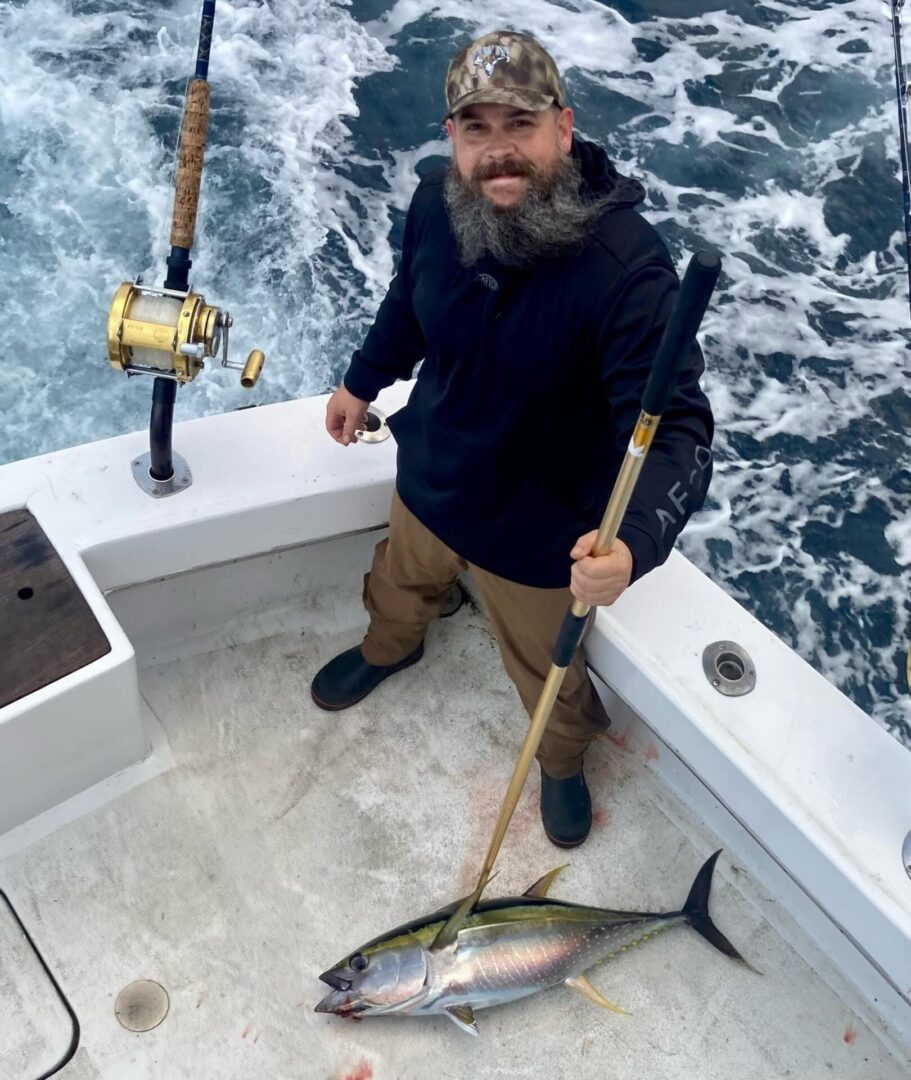 A fisherman stands on a boat holding a gaff with a freshly caught fish at his feet.