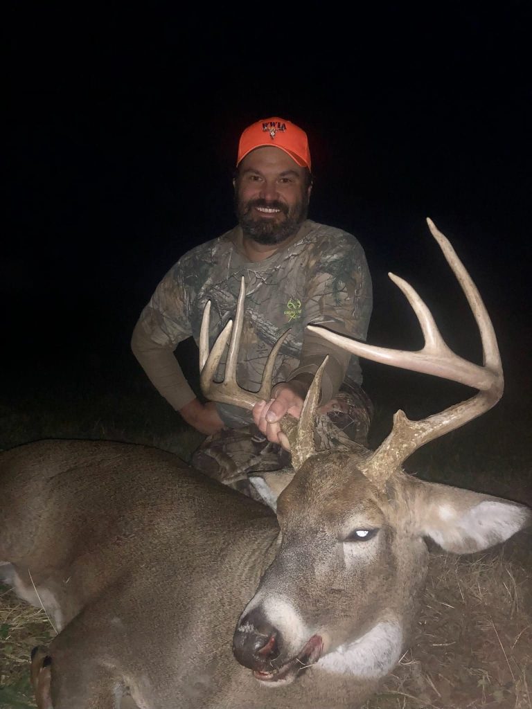 Hunter in camouflage posing at night with a large buck and impressive antlers, wearing an orange cap and smiling.